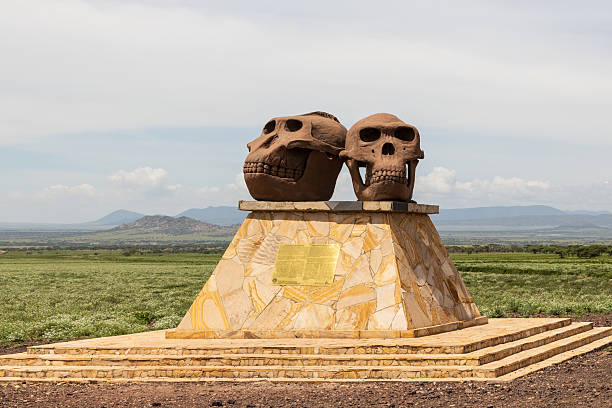 Tanzania - Serengeti - Olduvai Gorge - A striking monument featuring two large bronze hominid skulls sits atop a stone pyramid base on the open savannah plains near Olduvai Gorge, with the flat Serengeti landscape stretching to distant mountains. This landmark commemorates the groundbreaking paleoanthropological discoveries that made this region a crucial UNESCO site for understanding human evolution.