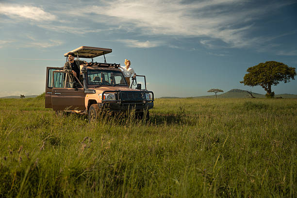 Low-angle view of a man and woman standing on the side steps of a safari vehicle with both doors open in the vast green grasslands of Serengeti National Park. Each looks in a different direction across the savannah landscape, with a lone tree visible in the distance under warm daylight. The image captures exploration, partnership, and the spirit of African safari adventure.